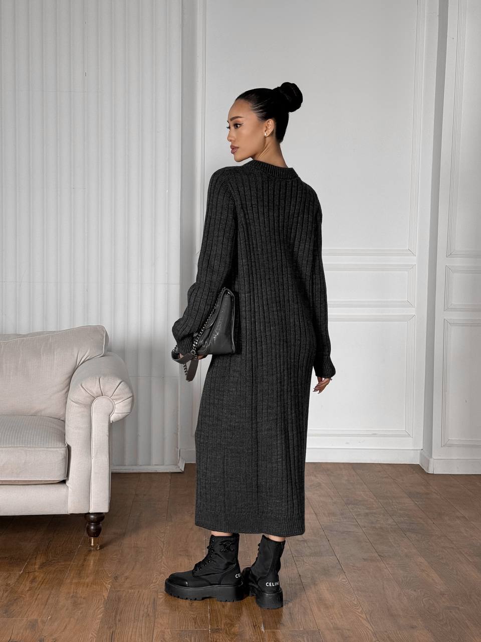 Woman in a long black ribbed sweater and gray skirt standing in a room with a white wall and beige sofa.