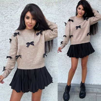 Woman wearing a beige sweater with black bows and a black pleated skirt against a white wall.