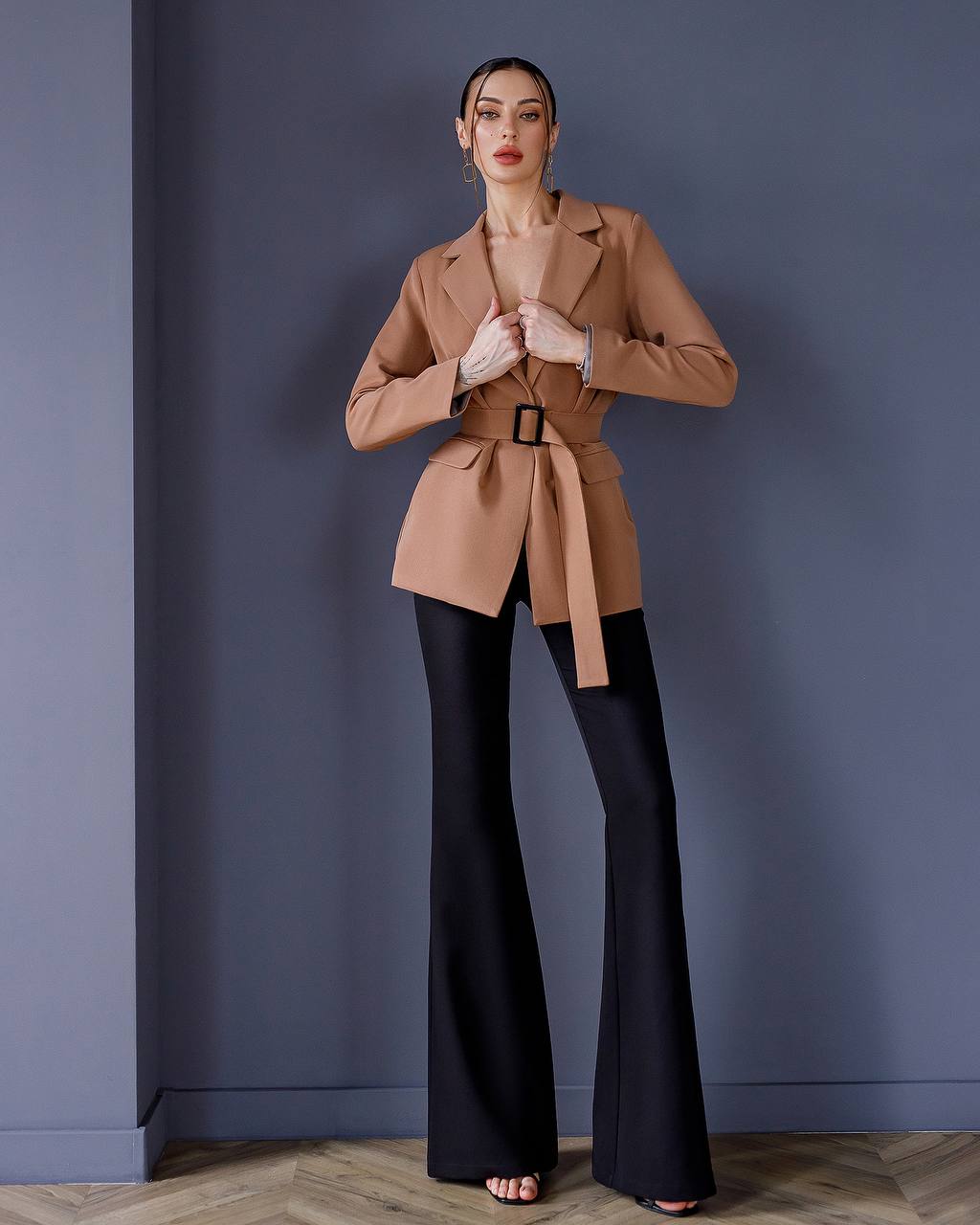 Woman wearing a beige trench coat and black pants against a blue wall.