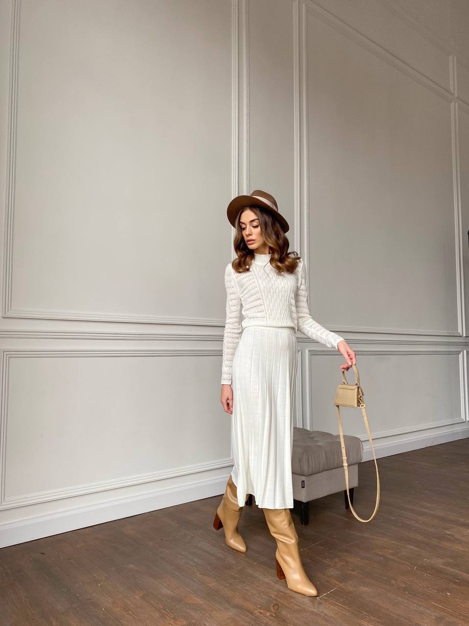 Woman in a white outfit with beige boots and a hat, holding a beige handbag against a neutral wall.