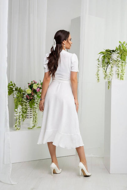 Woman in a white dress standing in a minimalistic room with white walls and a plant.