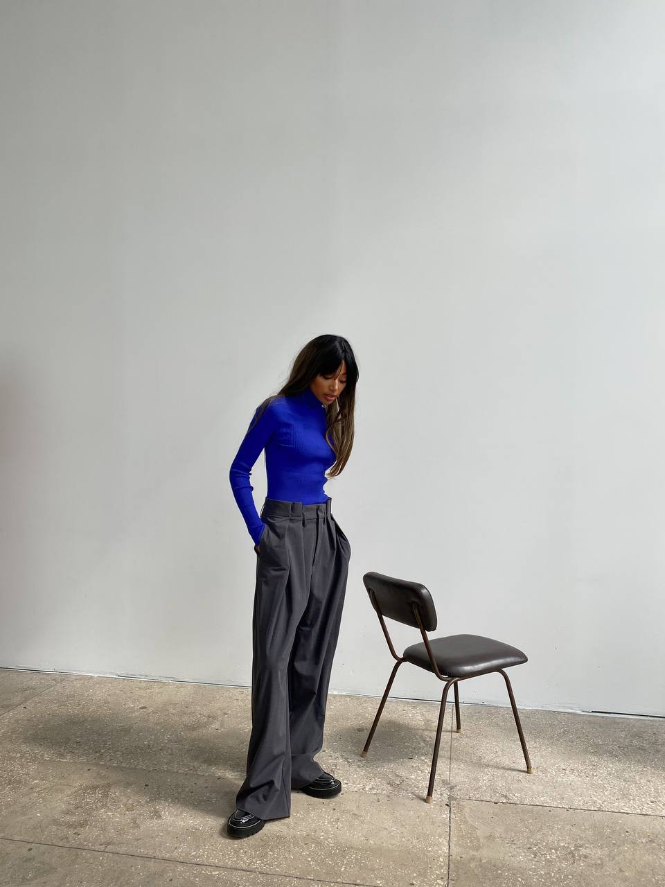 Person wearing a blue top and gray pants standing next to a black chair against a white wall.