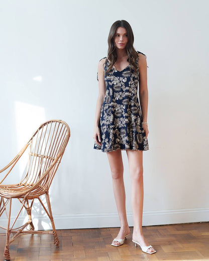 Woman wearing a navy floral dress standing next to a wicker chair against a white wall.