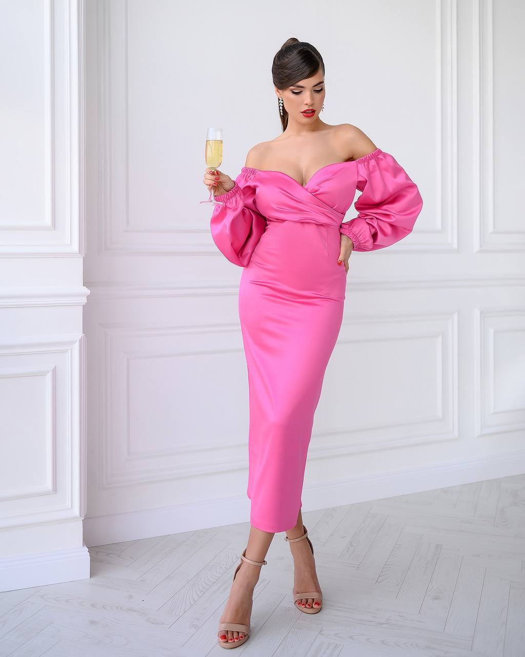 Woman in a pink off-shoulder dress holding a glass against a white wall.