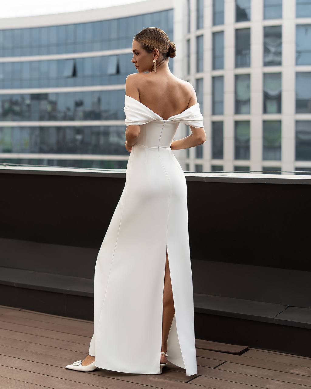 Woman in a white off-shoulder gown with a high slit standing on a rooftop with cityscape background