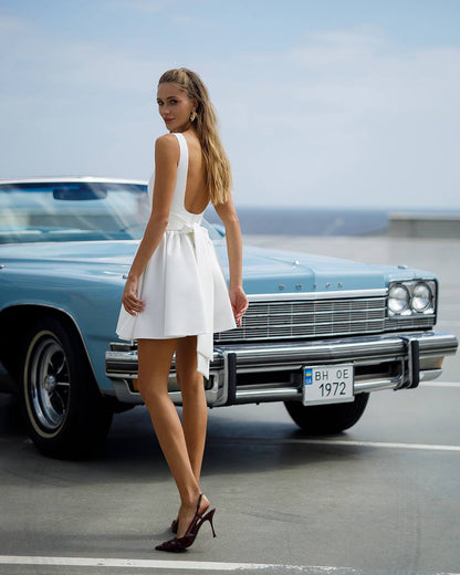 Woman in a white dress standing next to a classic car on a road.