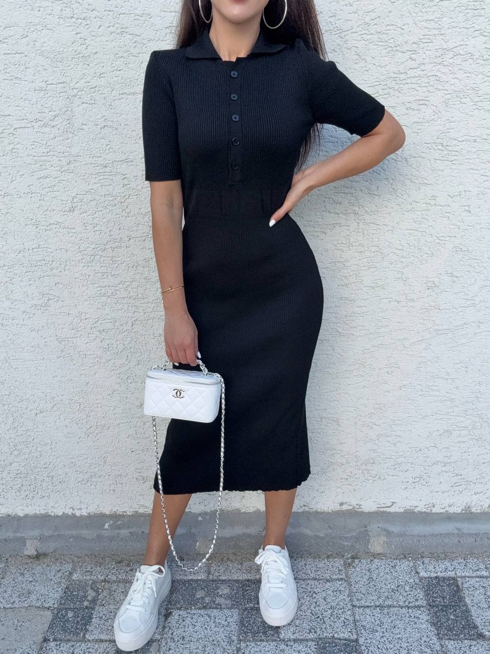Person wearing a black dress with a white handbag against a light gray wall.