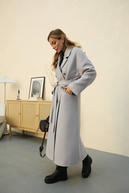Woman wearing a long gray coat in a room with a wooden cabinet and lamp.