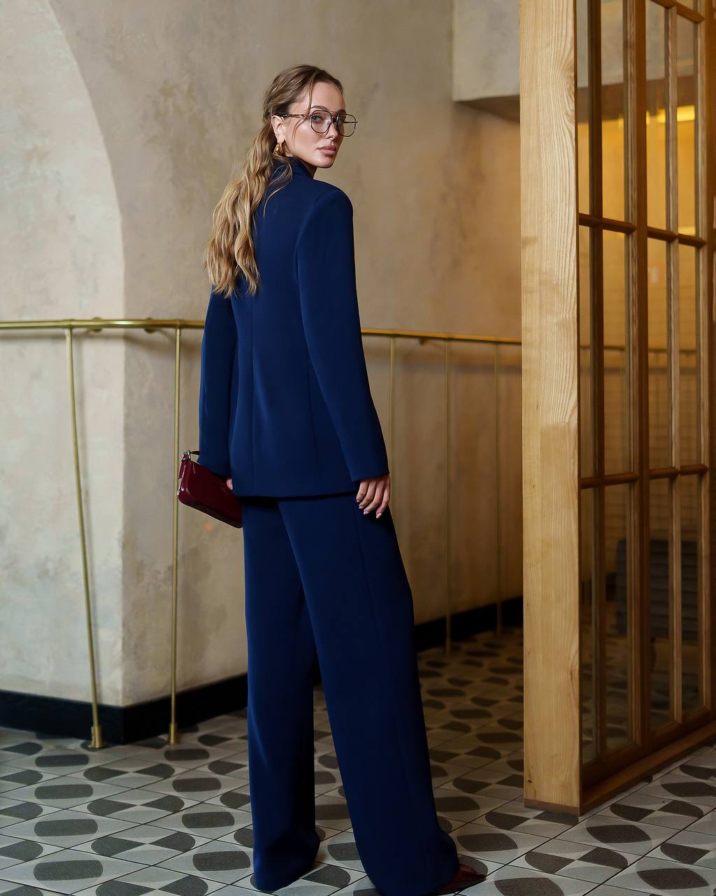 Woman in a blue suit standing in an elegant interior setting with a wooden door and patterned floor.