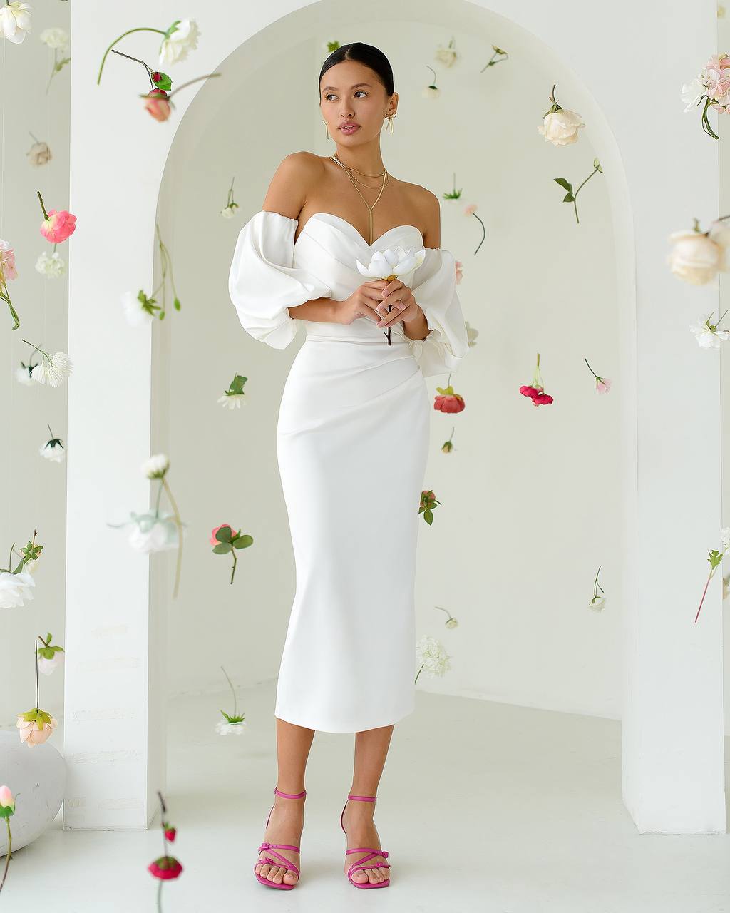 Woman in a white off-shoulder dress standing against a floral decorated wall.