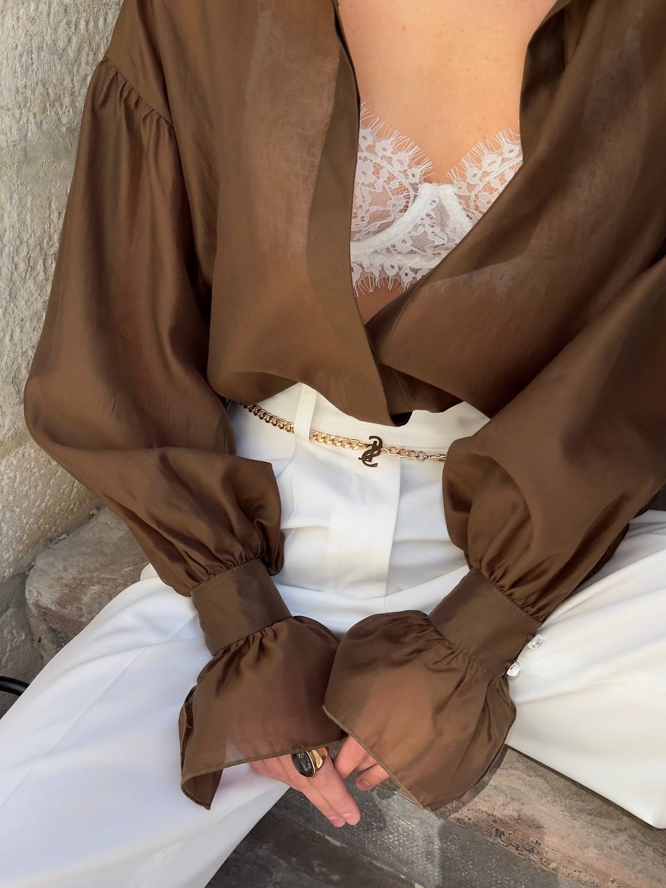 Brown blouse with long sleeves and white pants against a stone wall.