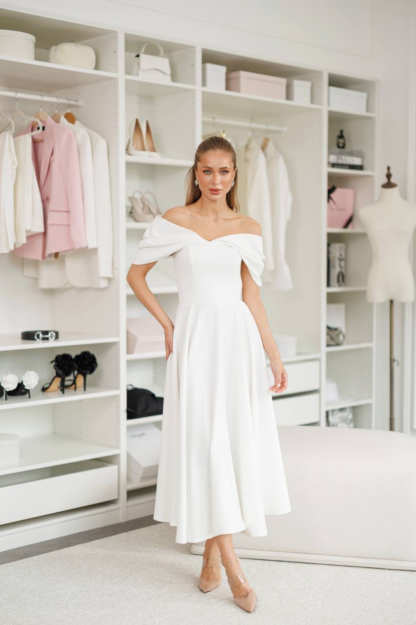 Woman wearing a white off-shoulder dress in a room with shelves and clothing.