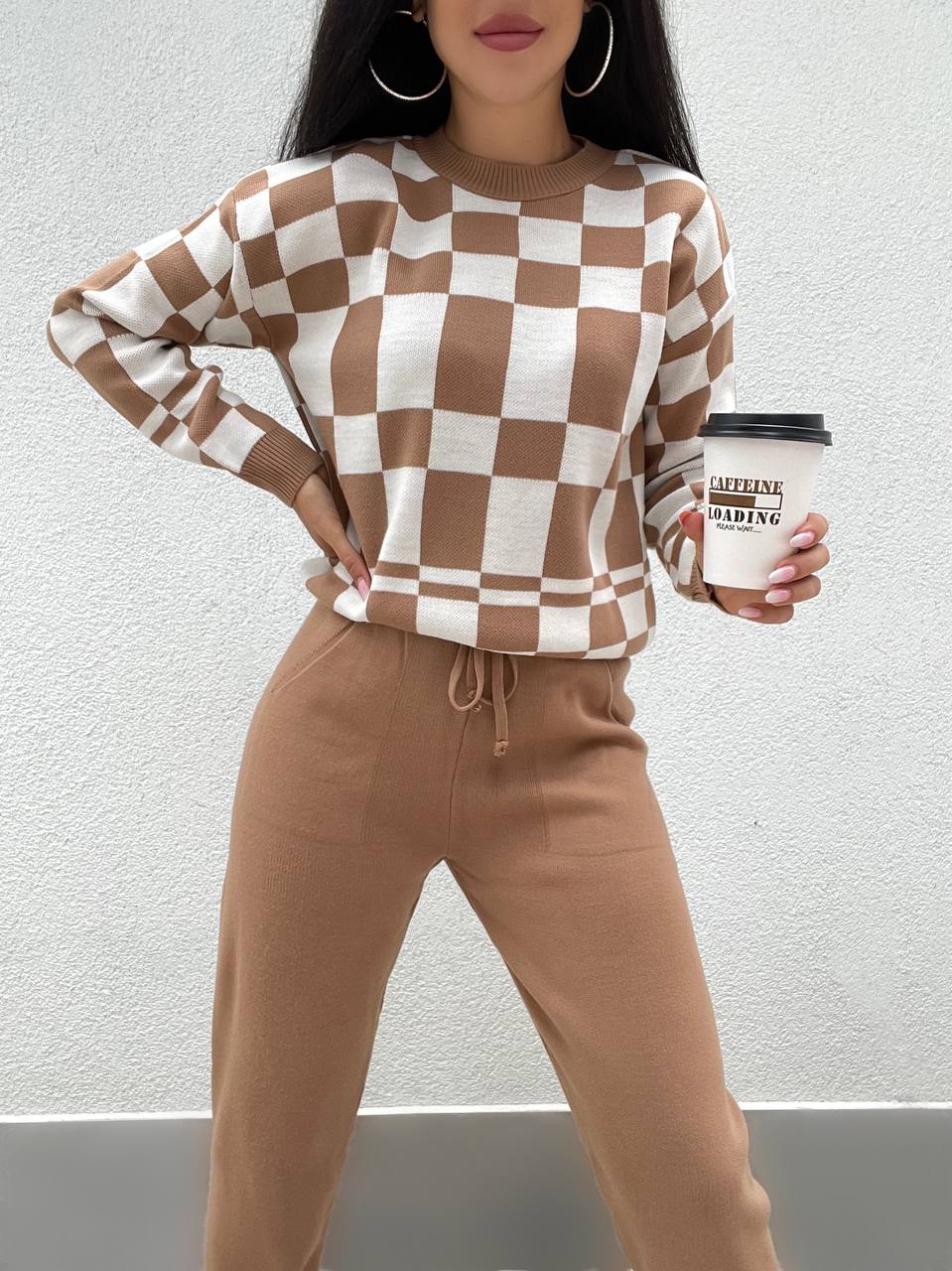 Person wearing a checkered brown and white sweater and beige pants, holding a coffee cup.