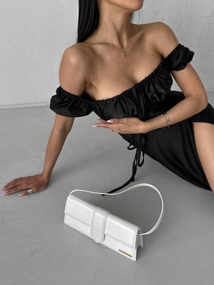 Woman in black off-shoulder dress with a white handbag on a gray floor.
