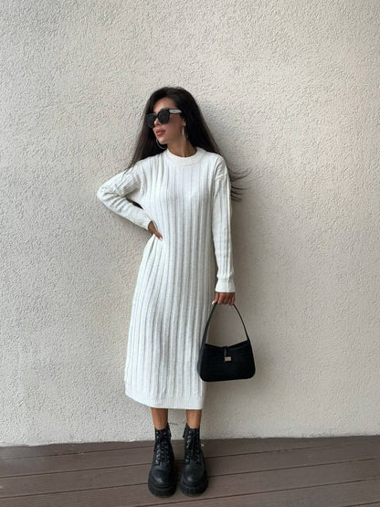 Woman wearing a white ribbed knit dress with black boots and a black handbag against a plain wall.