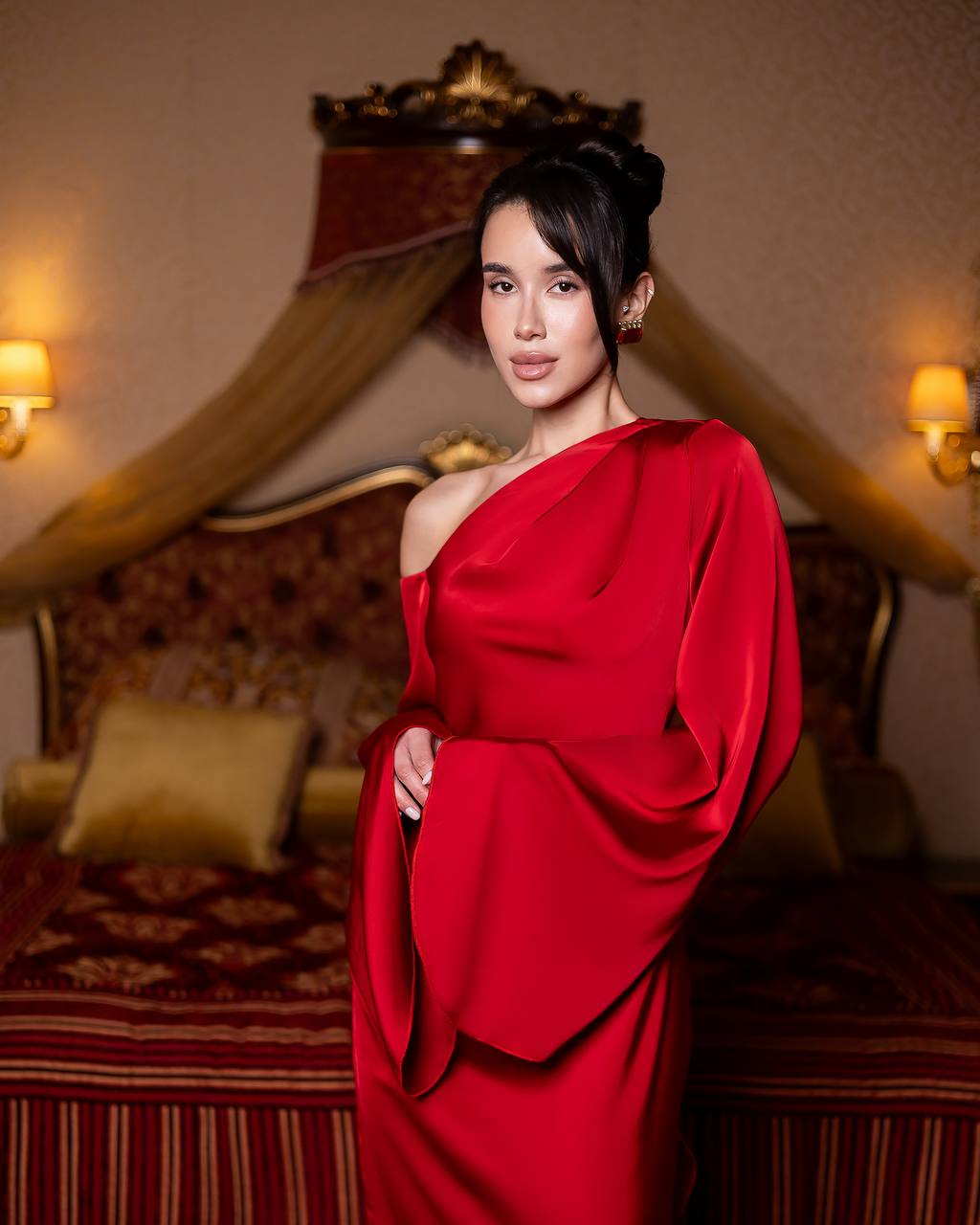 A woman wearing a vibrant red one-shoulder dress with dramatic draped sleeves, standing in a warmly lit, luxurious bedroom setting with a canopy bed in the background.
