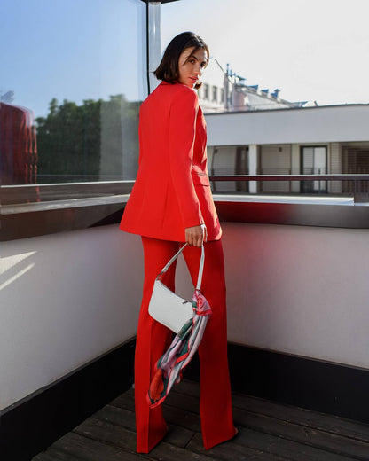 Woman in a red suit holding a white handbag on a balcony.