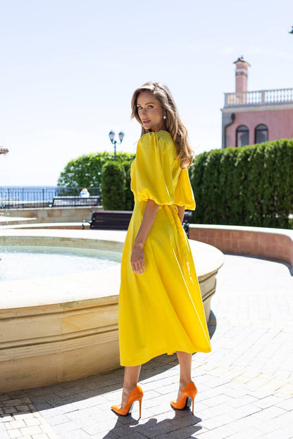 Woman in a yellow dress standing by a pool with greenery and a building in the background