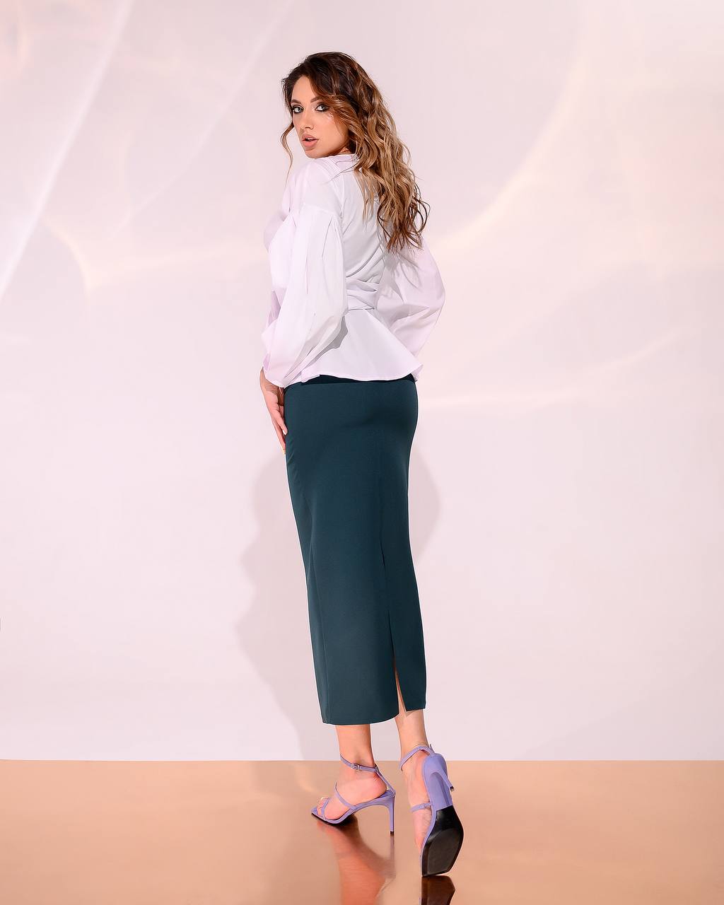 Woman wearing a white blouse and teal pants against a plain background