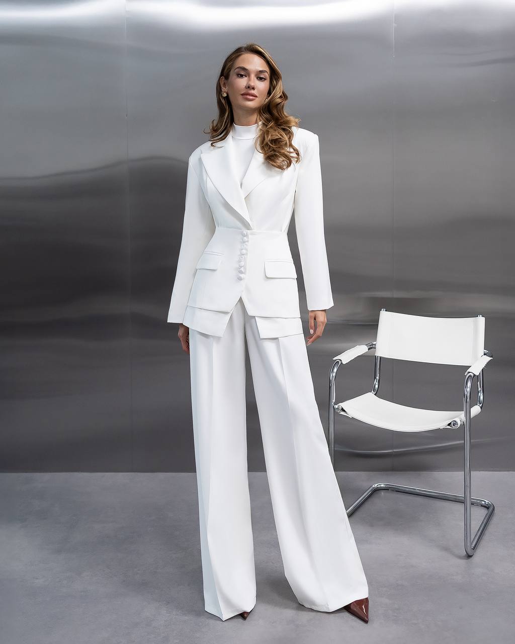 Woman in a white suit standing next to a white chair against a gray background