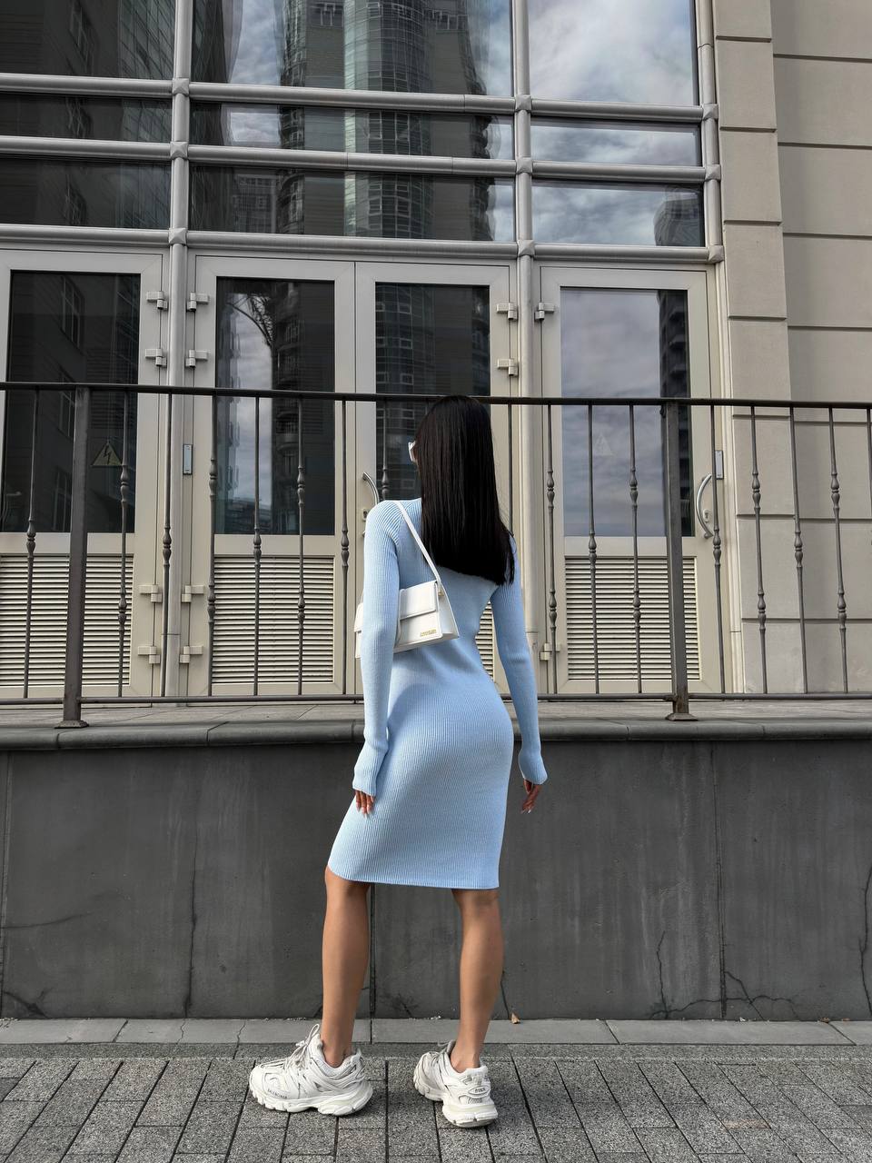 Person wearing a light blue dress standing on a city street with a modern building in the background.