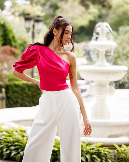 Woman in a bright pink top and white pants standing outdoors with a fountain in the background