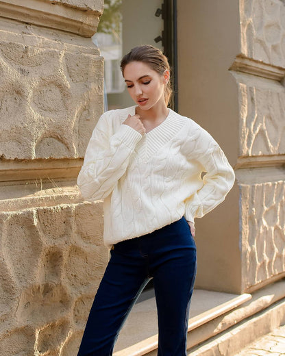 Woman wearing a white cable knit sweater and dark blue pants standing against a stone wall.