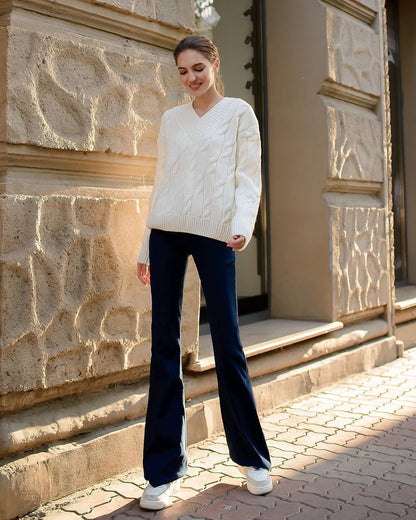 Woman wearing a white cable knit sweater and dark blue jeans standing in front of a stone building.