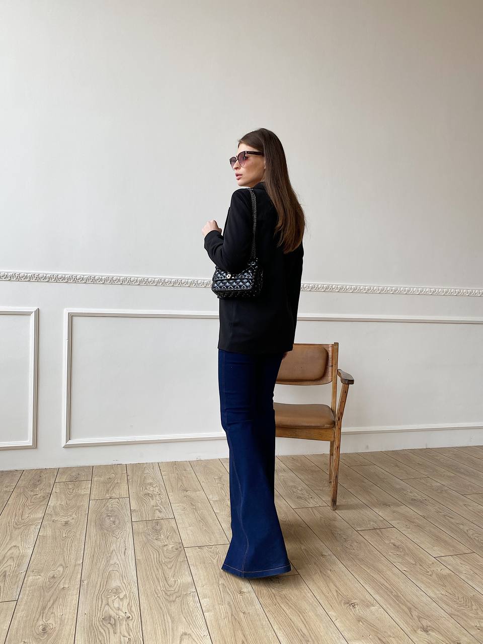 Woman in black sweater and blue jeans standing in a room with wooden floor and white walls.