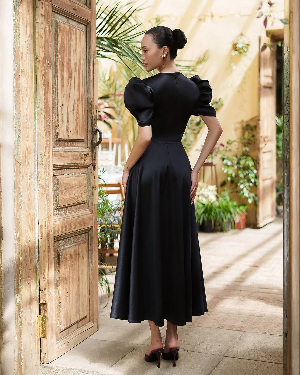 Woman in a black dress standing in a sunlit outdoor area with wooden doors and plants.