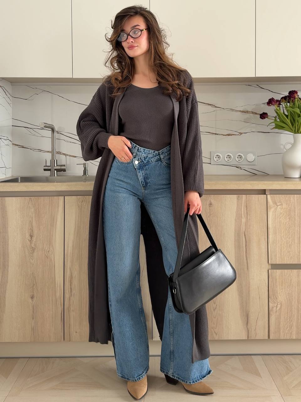 Woman standing in a kitchen wearing a long dark coat, black top, blue jeans, and holding a black handbag.