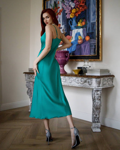 Woman in a teal dress standing in an elegant room with a decorative table and colorful painting.