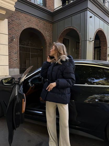 Woman in a navy puffer jacket standing next to a black car in an urban setting.