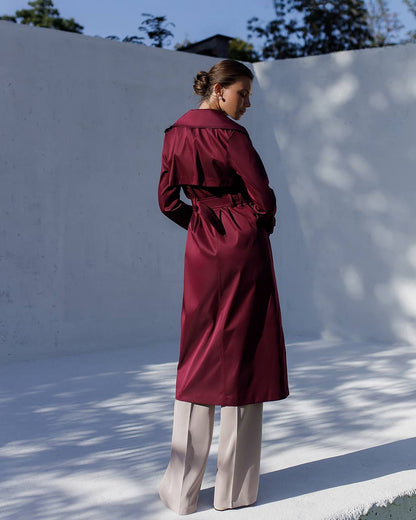 Person wearing a burgundy trench coat standing against a white wall with trees in the background