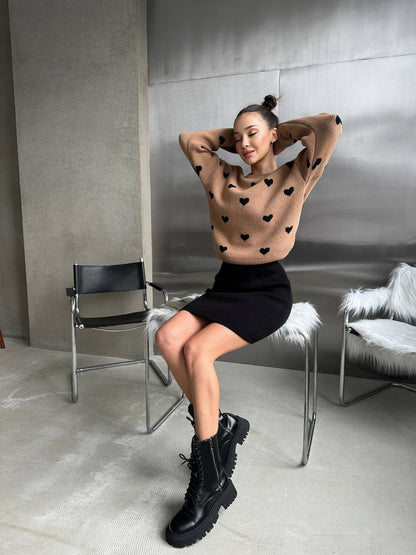 Woman wearing a heart-patterned sweater and black skirt sitting on a chair in a modern interior setting.