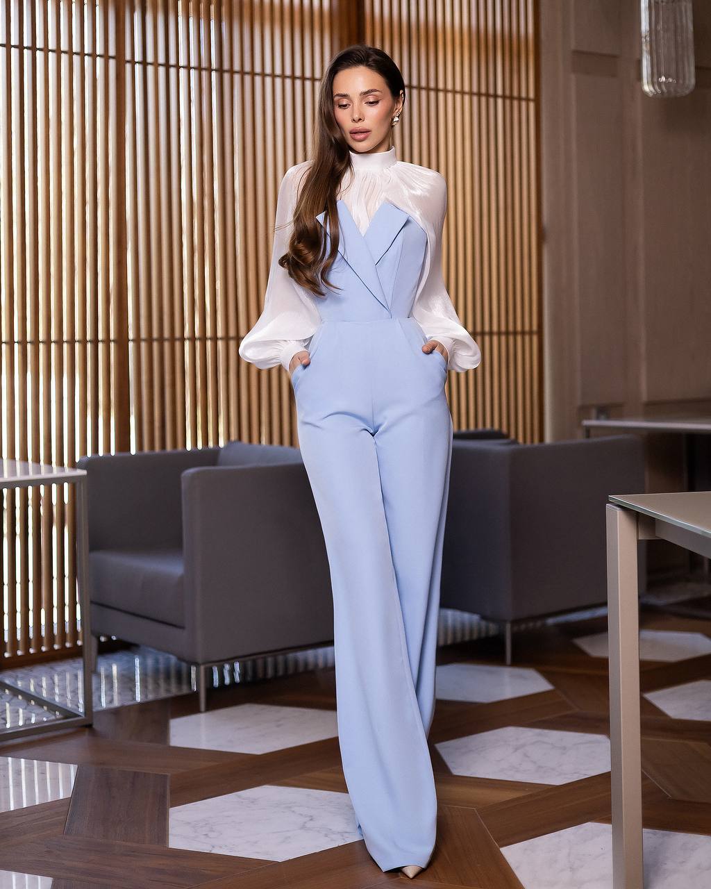 Woman in a light blue suit and white blouse standing in an indoor setting with wooden flooring and furniture.