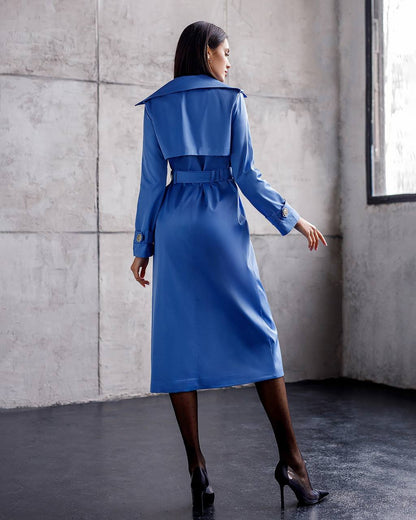 Woman wearing a blue trench coat standing against a concrete wall.