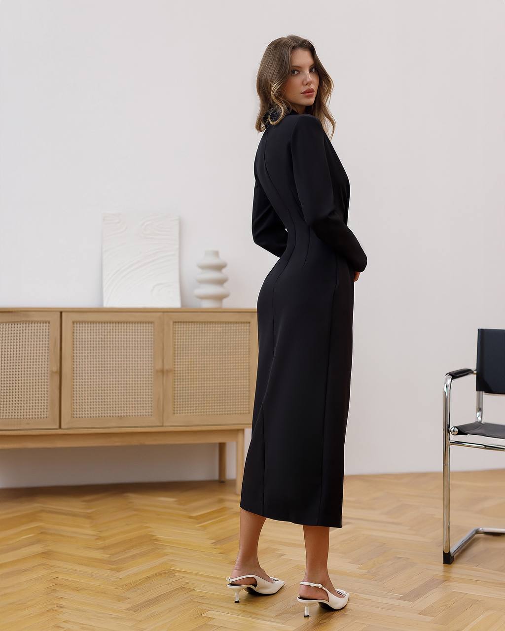 Woman in a black dress standing in a room with wooden flooring and a light-colored wall.