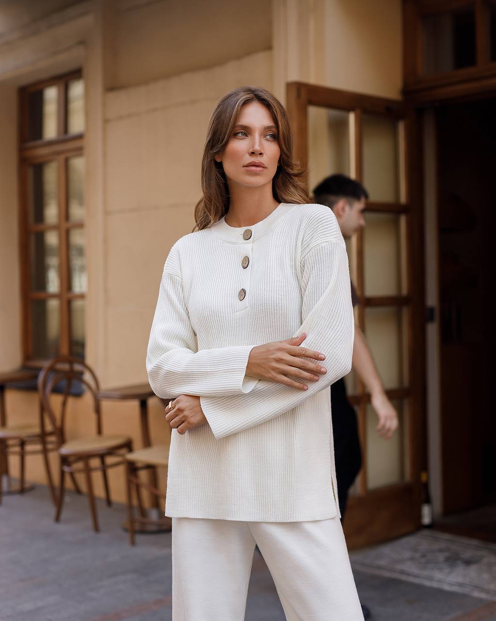 Woman in a white outfit standing in an indoor setting with wooden furniture.