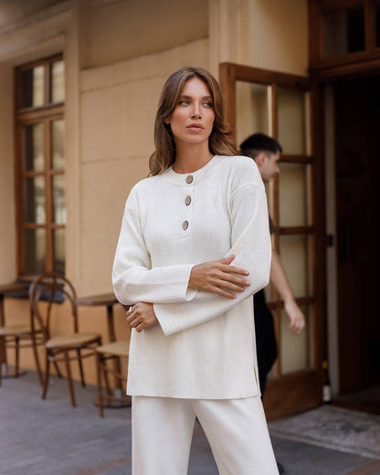 Woman in a white outfit standing in an indoor setting with wooden furniture.