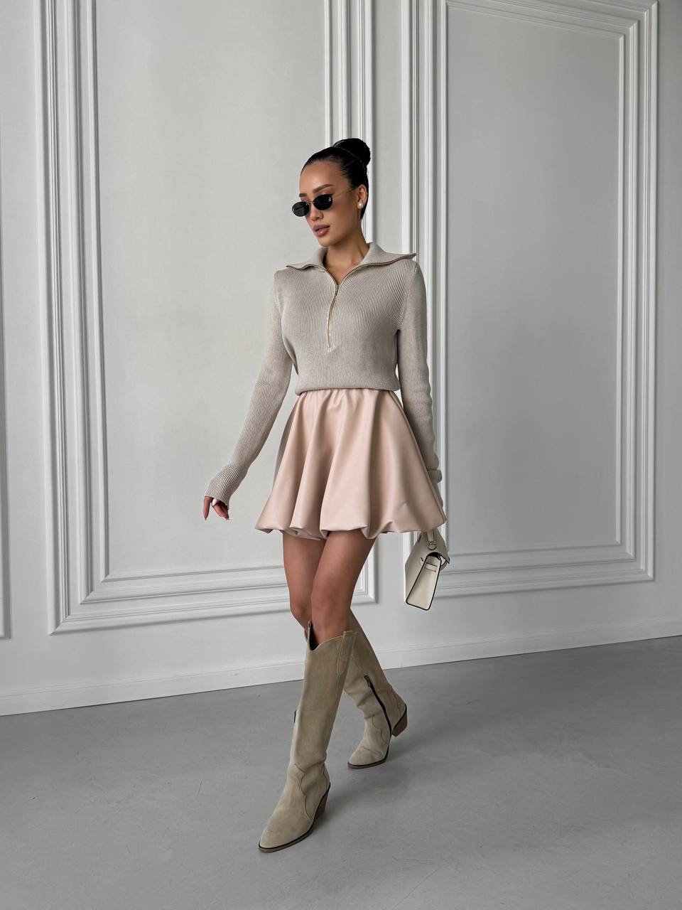 Woman wearing a beige sweater and skirt with knee-high boots against a gray wall.