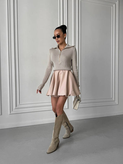 Woman wearing a beige sweater and skirt with knee-high boots against a gray wall.