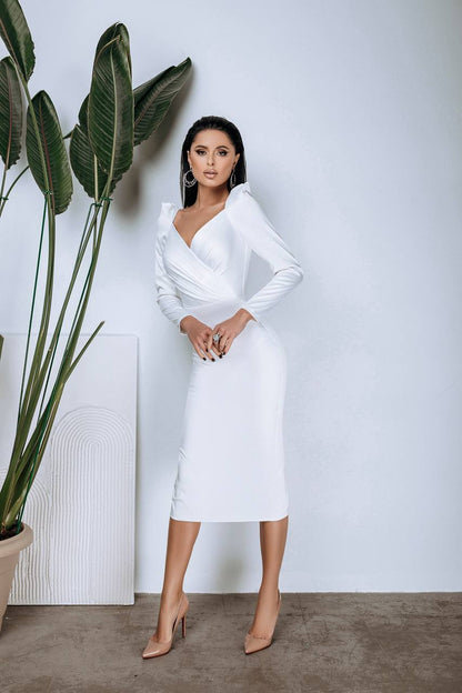 Woman in a white dress standing next to a plant against a plain wall