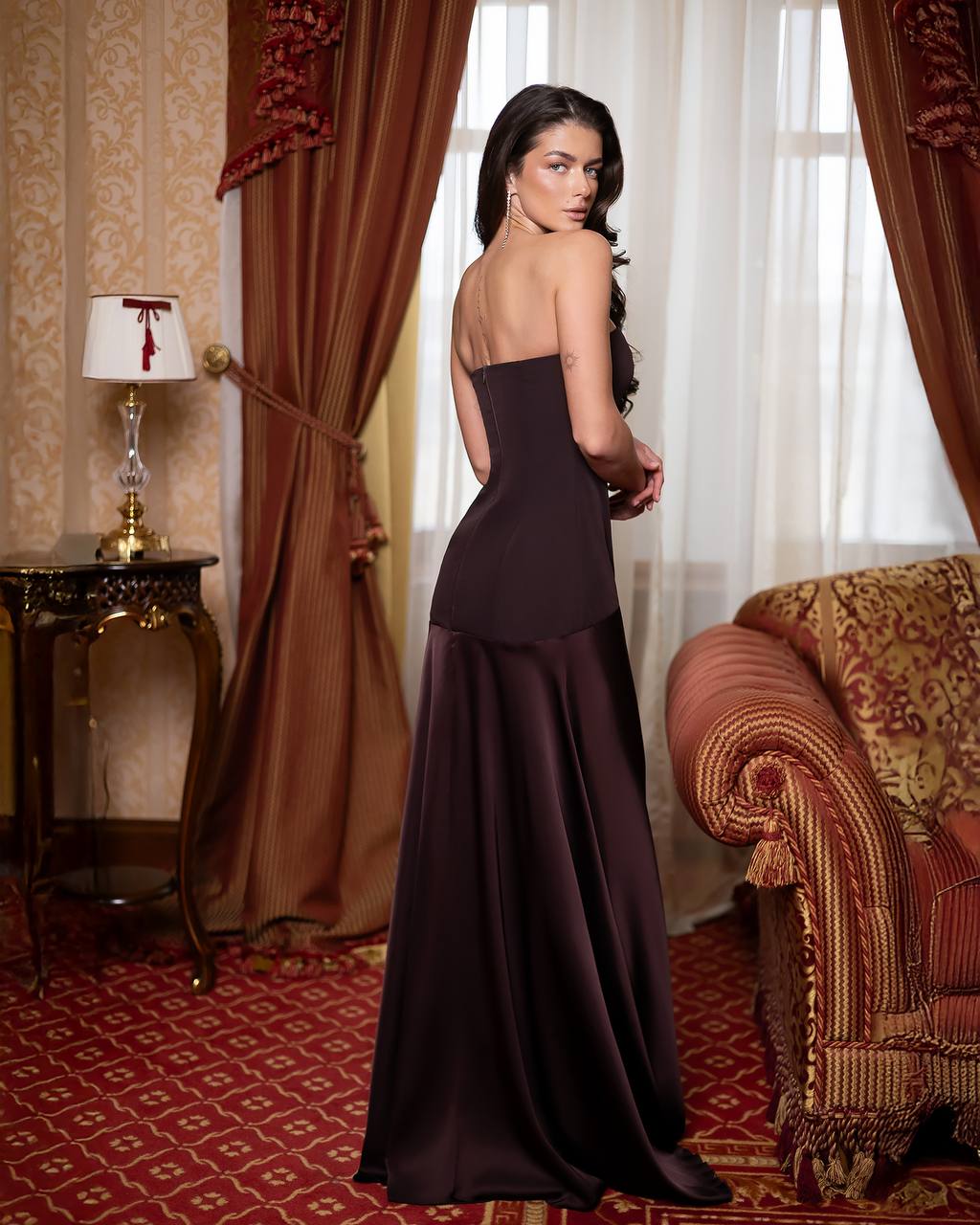 Woman in a dark purple evening gown standing in an opulent room with red and gold decor.