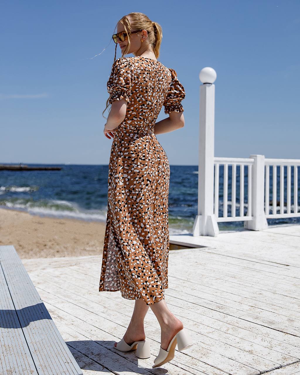 Woman in a patterned dress standing on a wooden deck by the beach.