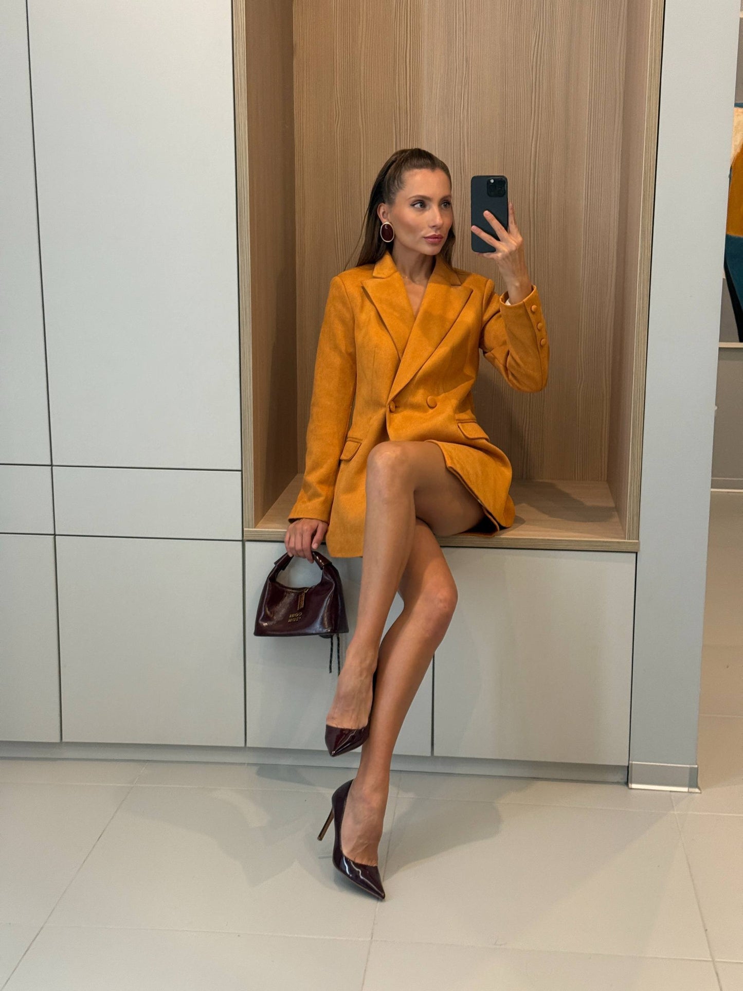 A woman takes a mirror selfie in a fitting room wearing a mustard yellow blazer dress, holding a dark burgundy handbag and wearing dark burgundy pointed-toe heels. She is seated on a built-in wooden bench against a white and wood cabinet background.