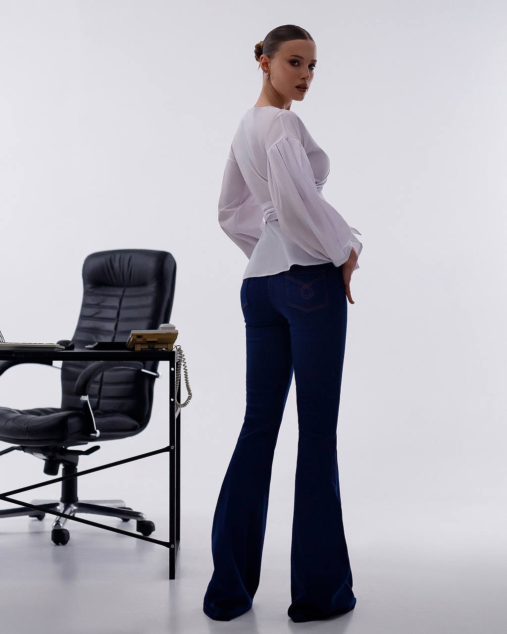 Woman in a white blouse and blue jeans standing next to a black office chair on a white background