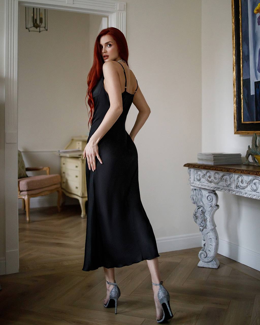 Woman in a black dress standing in an elegant room with wooden flooring and decorative furniture.
