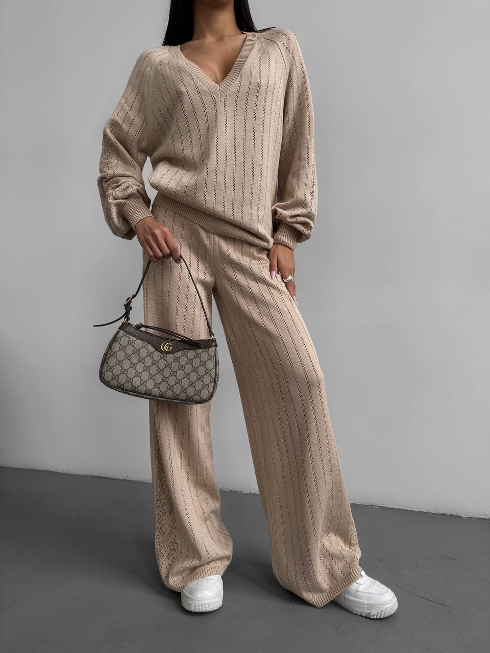 Person wearing a beige knit outfit holding a Gucci handbag against a gray background