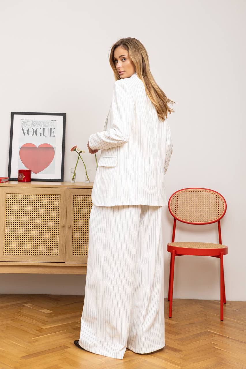 Woman in a white outfit standing in a room with a red chair and wooden cabinet.